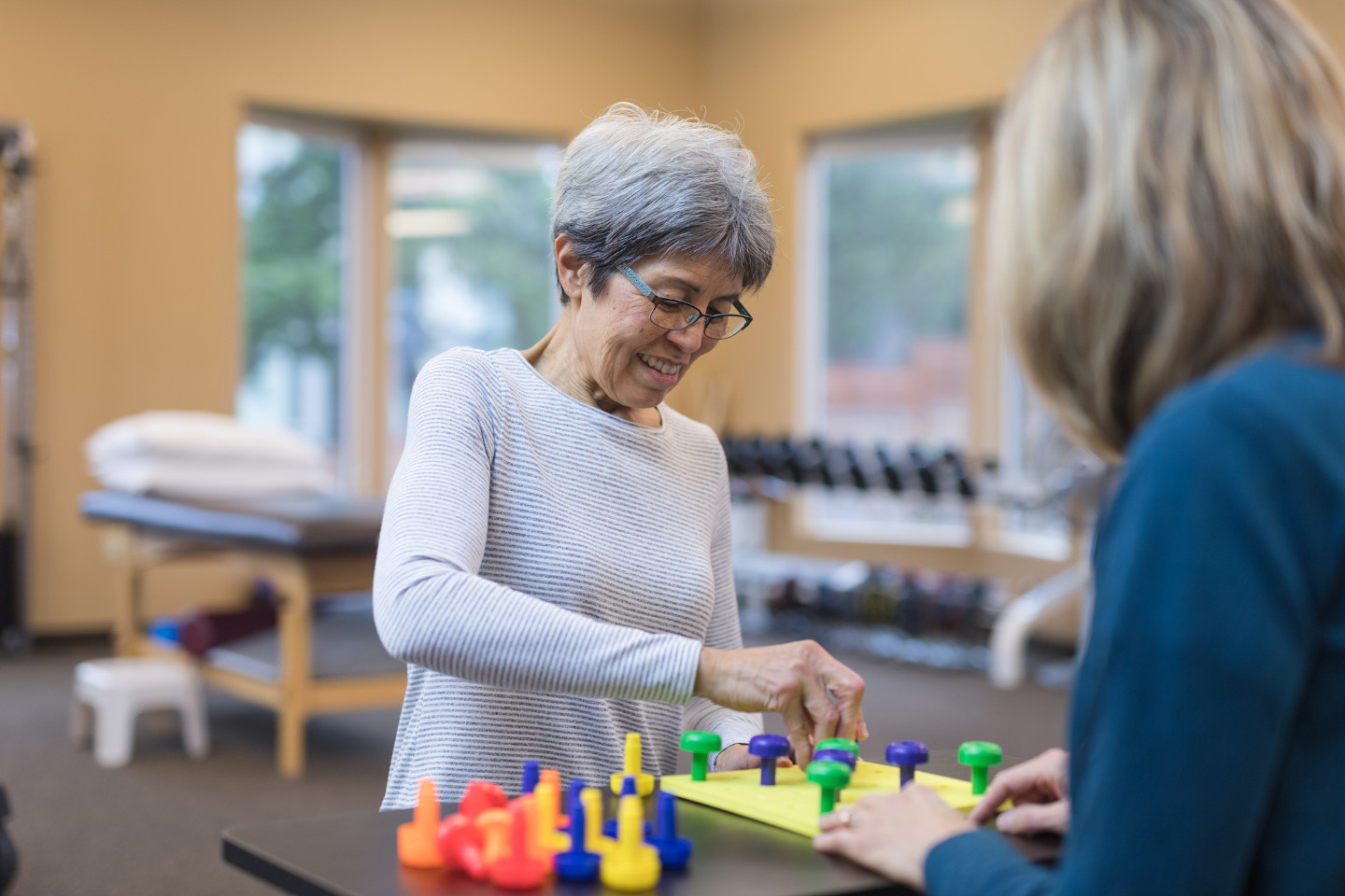 An occupational therapist works with a senior Caucasian woman They are seated at a table and they are doing a fun exercise that involves putting pegs into a plastic board.